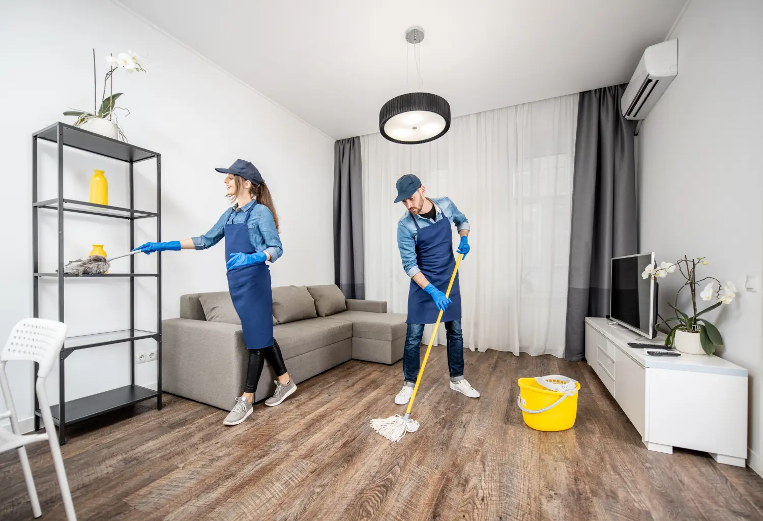 Professionelle Reinigungskräfte in blauer Uniform waschen den Boden und wischen Staub von den Möbeln im Wohnzimmer der Wohnung